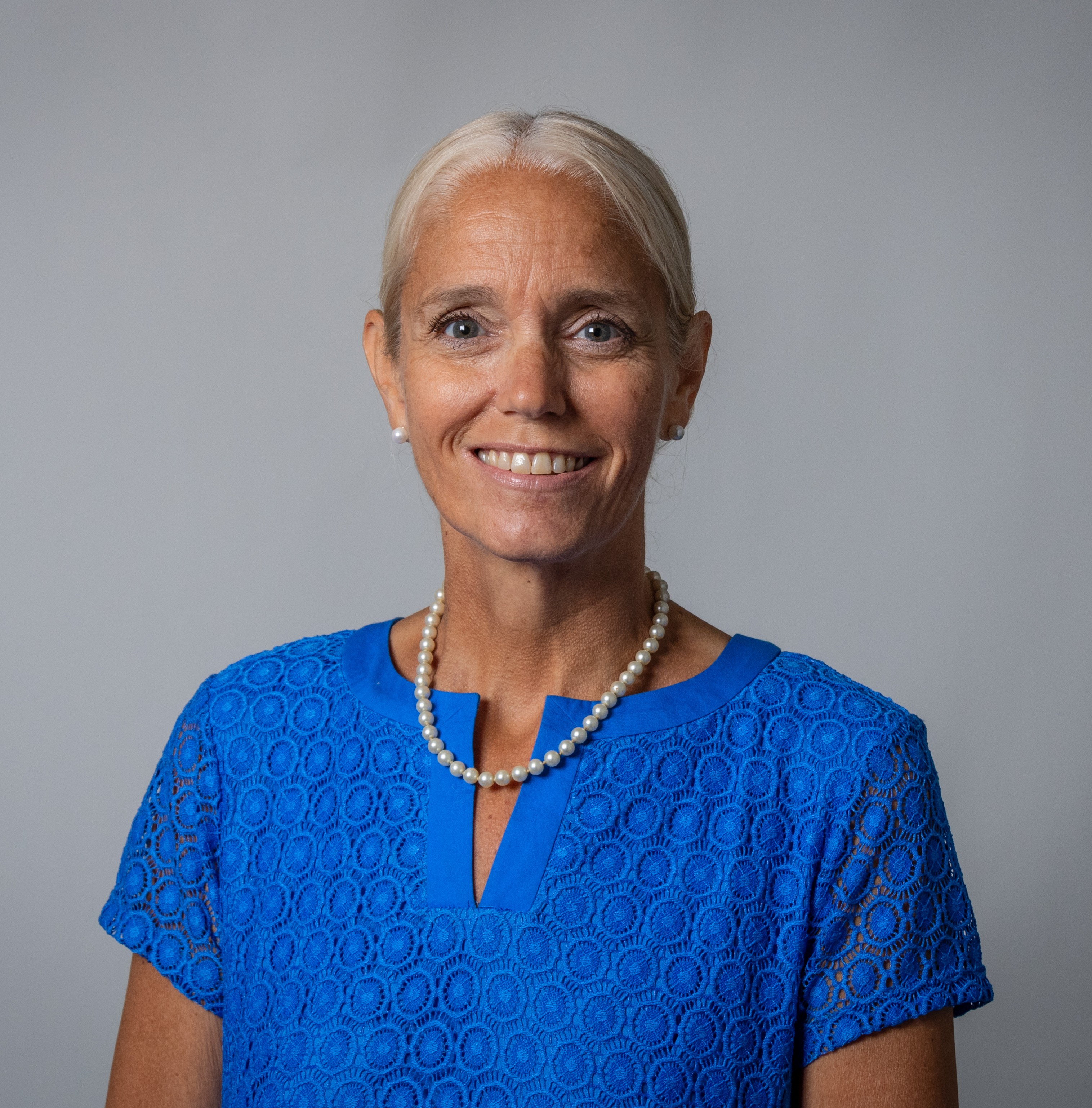 Stephanie Wanne smiling wearing a bright blue short sleeve lace top and pearl necklace against a plain gray background
