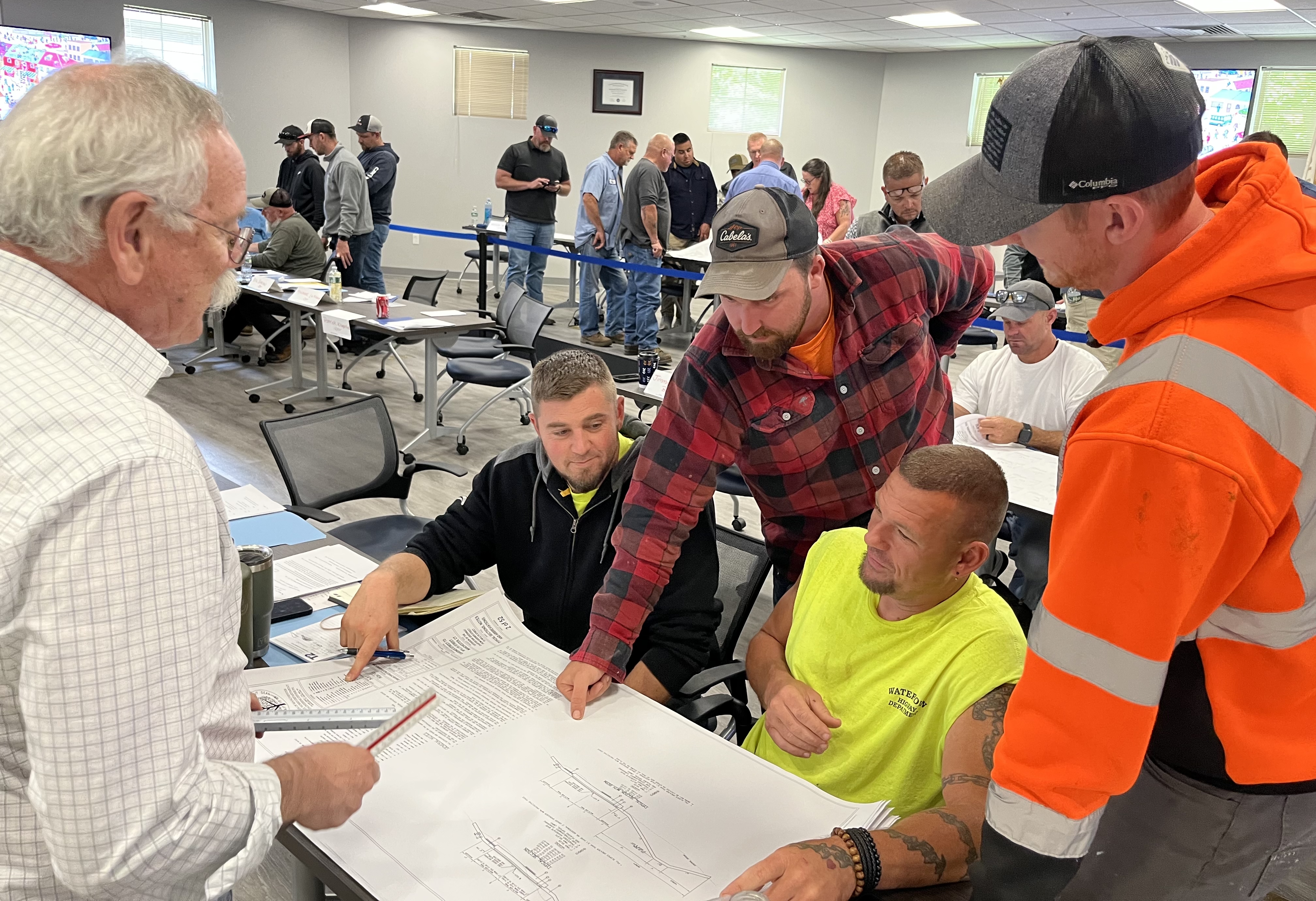 A group of public works employees collaborate around a large blueprint at a classroom table while an instructor looks on. More participants are seated and standing in the background during the training session.