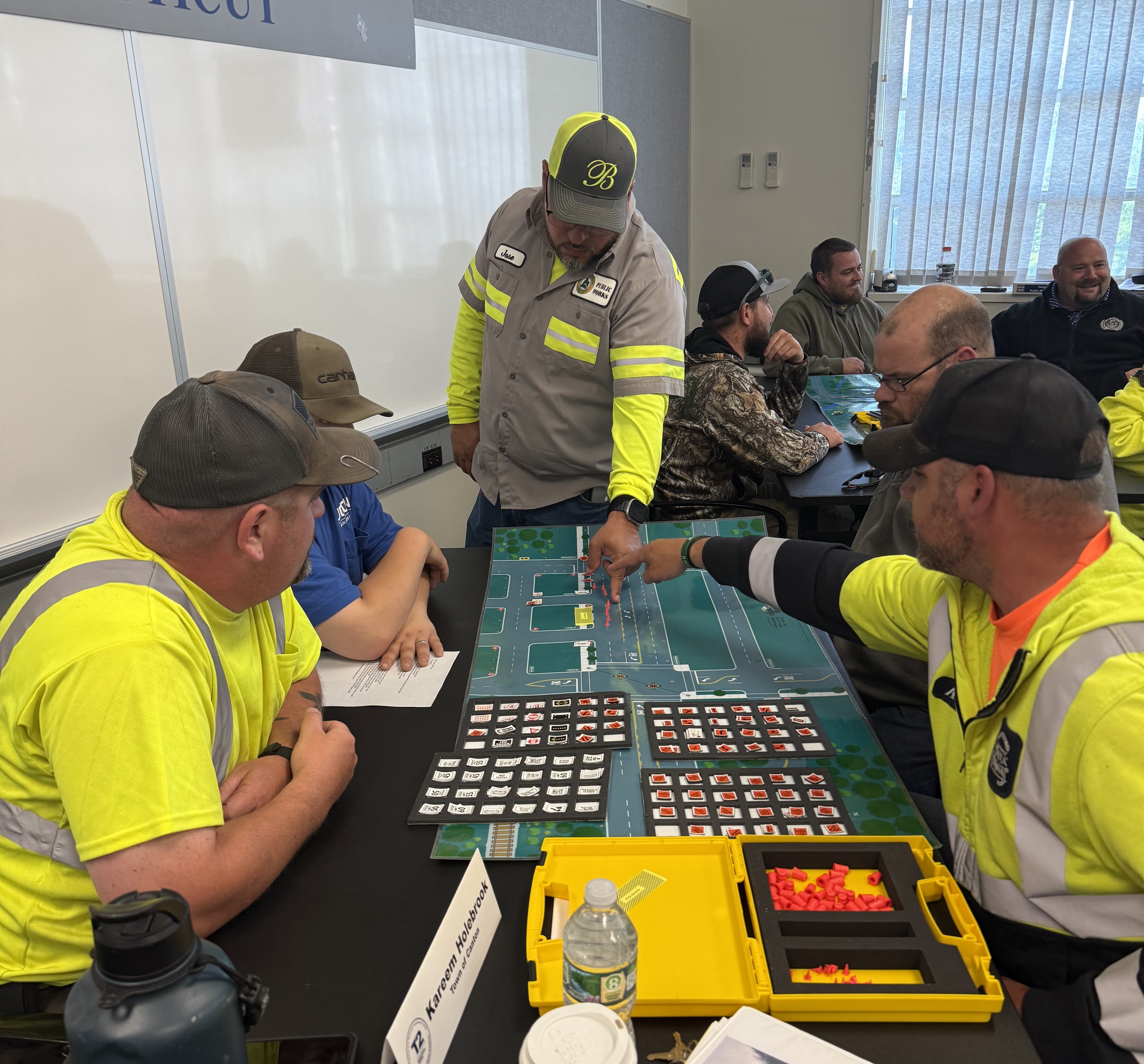 A group of public works professionals sits around a classroom table engaged in a hands-on training activity. Several men in high-visibility shirts and work uniforms are focused on a tabletop work zone simulation board covered with roadway diagrams, small traffic control pieces, and labeled cards. One participant points to the board while another observes closely, and others discuss strategies. Additional participants are seated at nearby tables, also working on similar activities. The room has whiteboards and windows in the background.